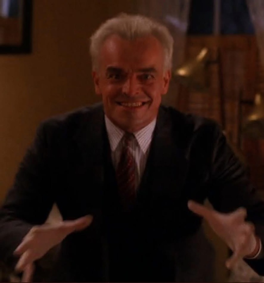 Mark Alford Bullying in Congress. A man that resembles Mark Alford in a dark suit with a white shirt and striped tie leans forward with a wide, unsettling grin and hands outstretched, creating an eerie and menacing expression.